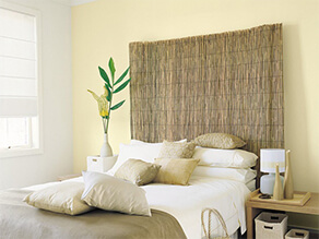Light yellow feature wall with bamboo bedhead and plant on bedside table with gold pillows